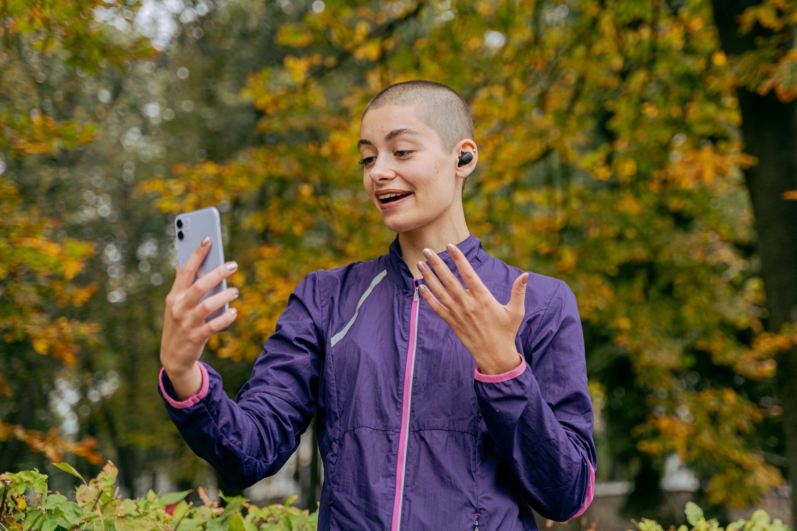 Junge Frau in violetter Jacke lächelt während eines Videoanrufs mit ihrem Smartphone in einem herbstlichen Park