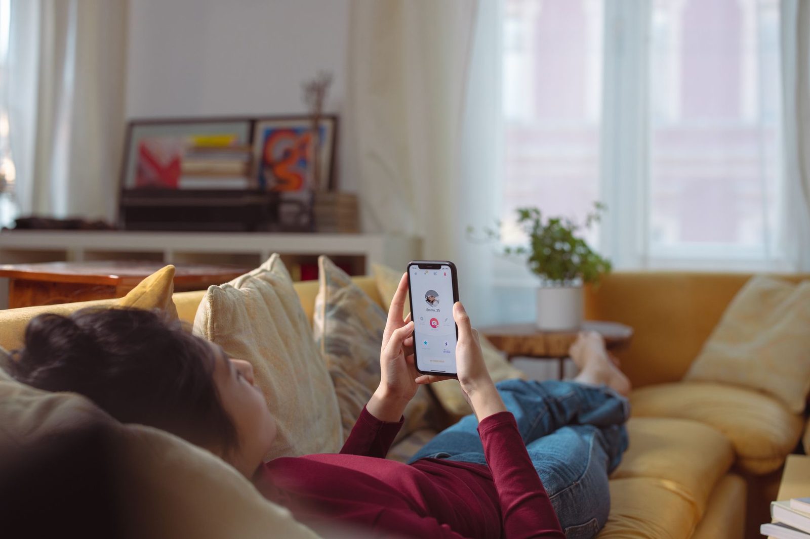 Junge Frau, die auf einem gelben Sofa liegt und eine Dating-App auf ihrem Smartphone nutzt