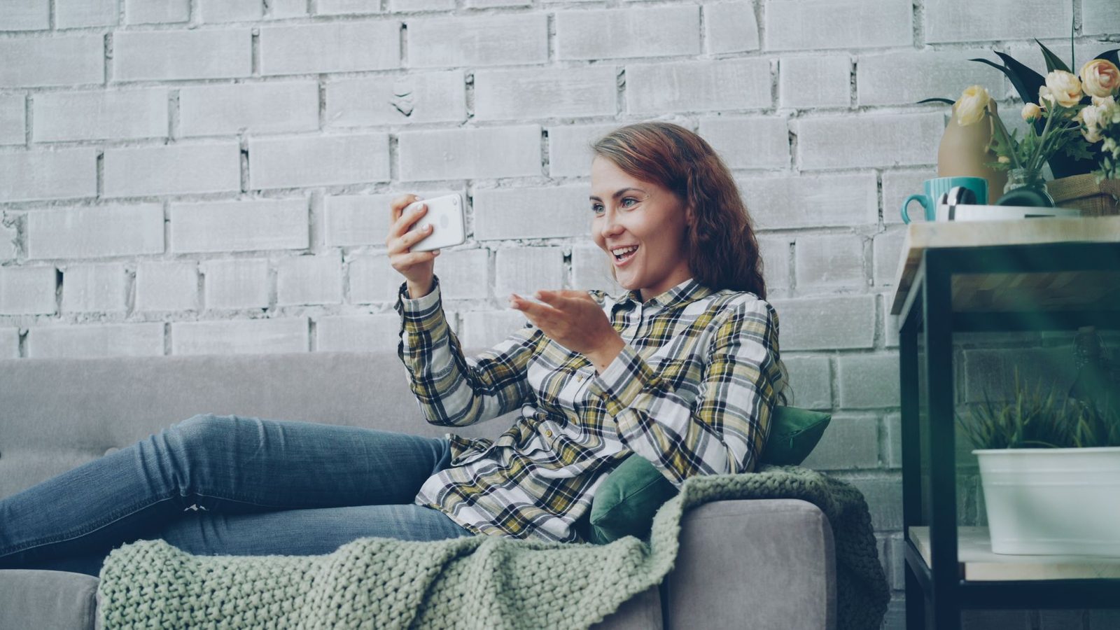 Lächelnde junge Frau auf einem Sofa, die in einem Wohnzimmer mit ihrem Smartphone einen Videoanruf führt