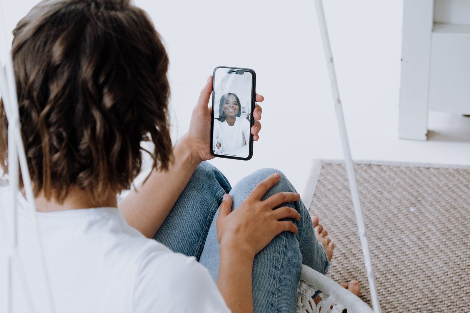 Sitzende Frau mit Smartphone in der Hand während eines Videoanrufs mit einer anderen lächelnden Frau auf dem Bildschirm