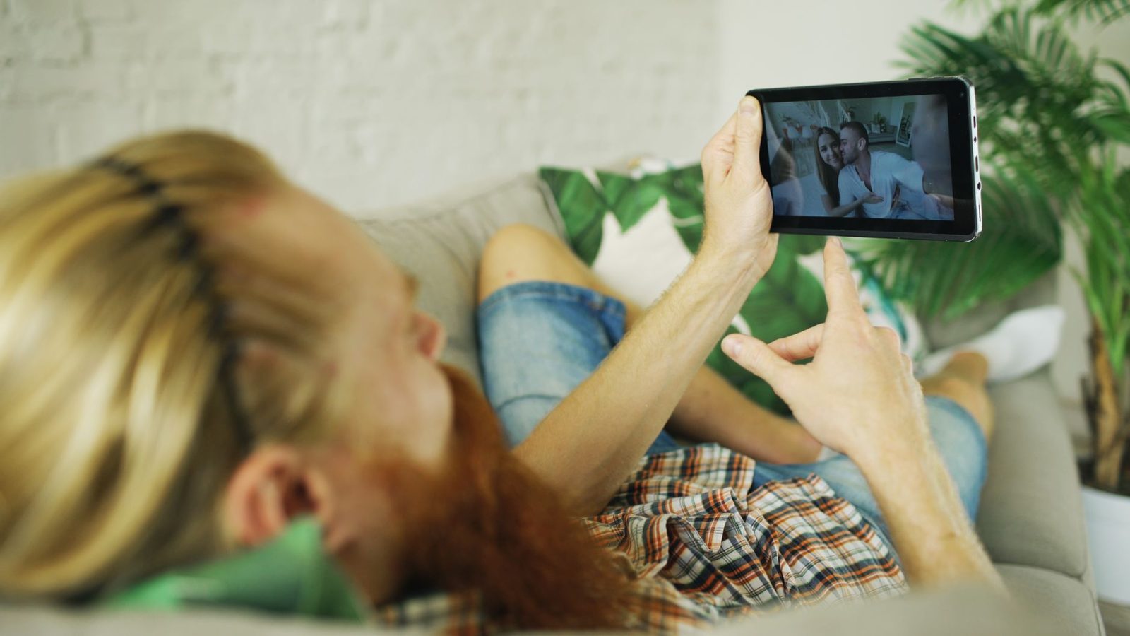 Man liggend op een bank met een tablet waarop een videogesprek met een koppel te zien is