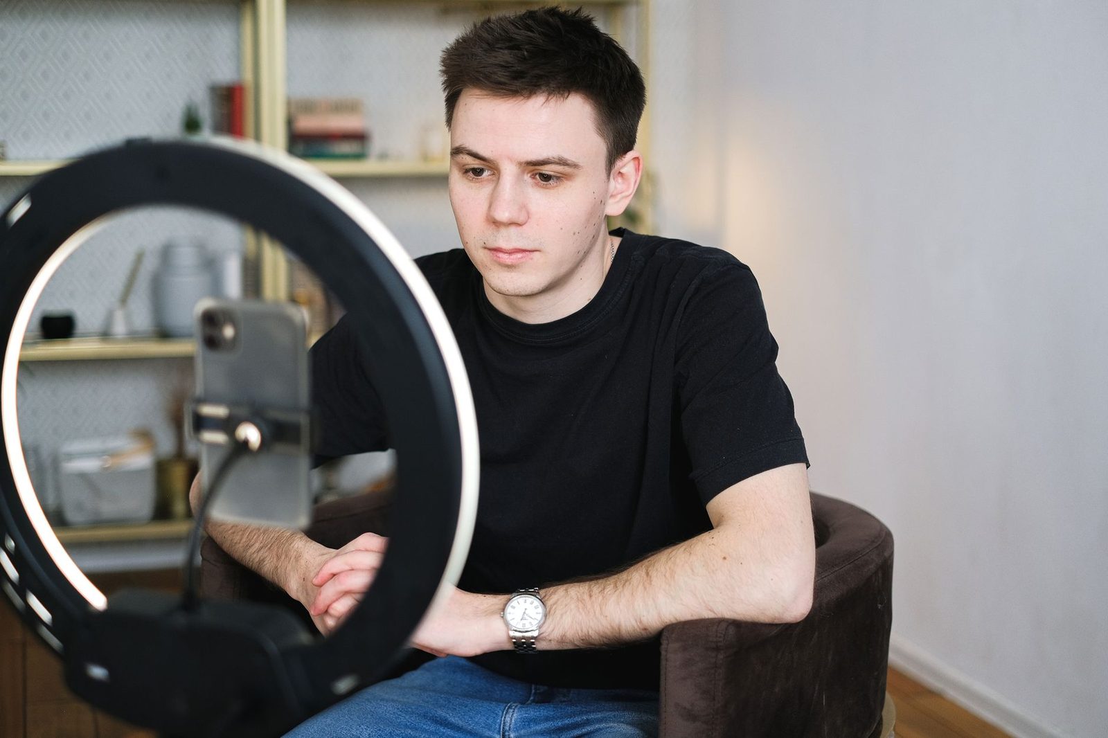 Junger Mann sitzt vor einem Smartphone auf einem Stativ und einem Ringlicht, beim Videochat zu Hause
