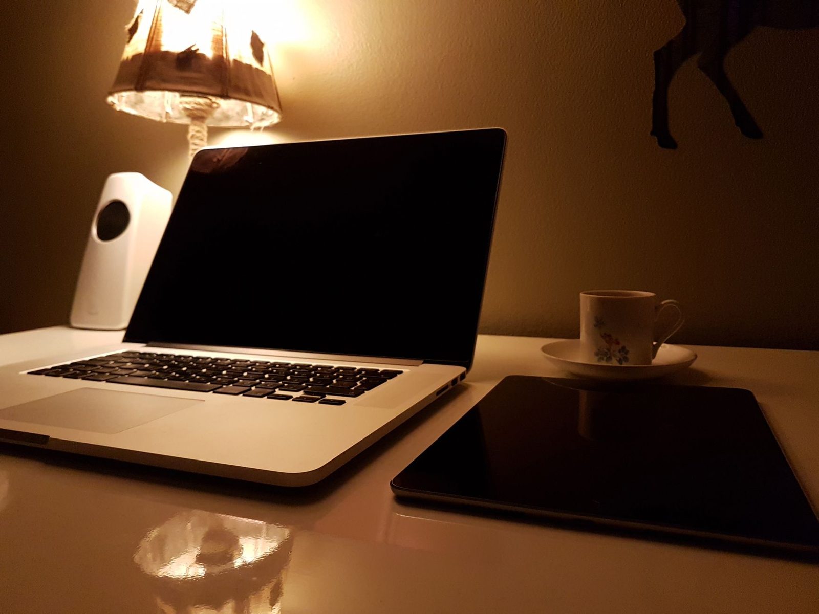 Aufgeklappter Laptop, Tablet und Kaffeetasse auf einem weißen Schreibtisch, beleuchtet von einer Lampe
