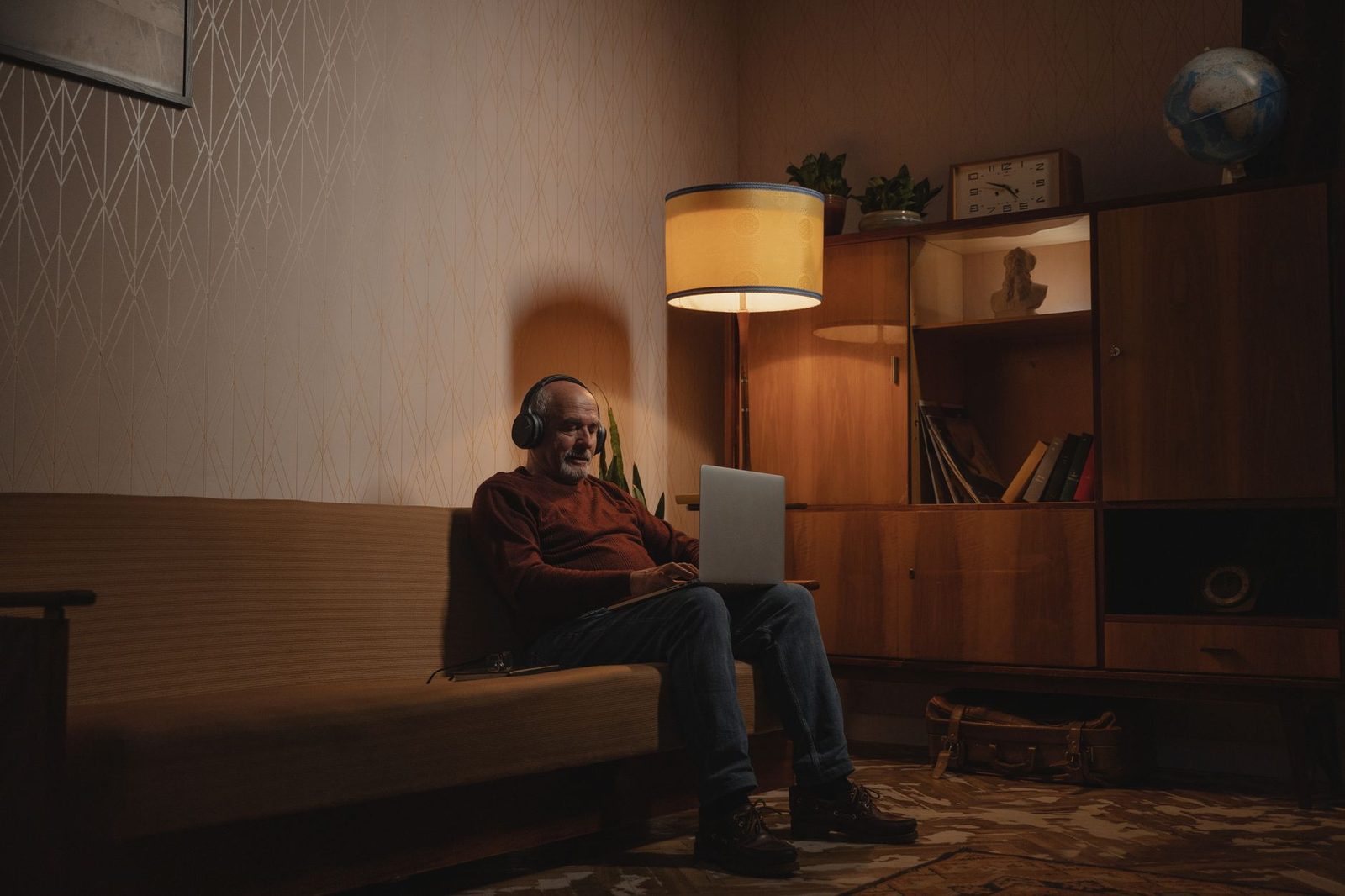 Hombre con auriculares sentado en un sofá, con el portátil sobre las rodillas, en un salón iluminado por una lámpara