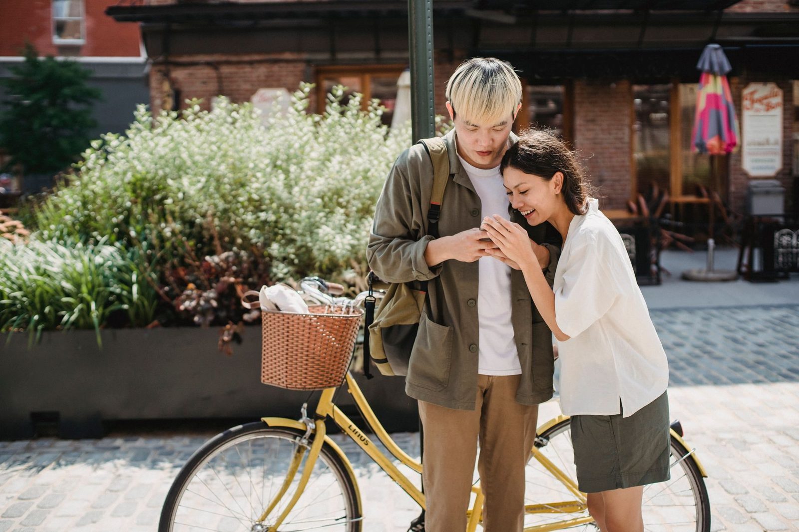 Una pareja joven sonriente mira junta un smartphone en una calle adoquinada, junto a una bicicleta amarilla