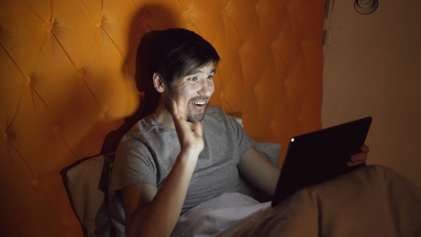 Lächelnder Mann, der abends im Bett liegt und vor einem Tablet winkt
