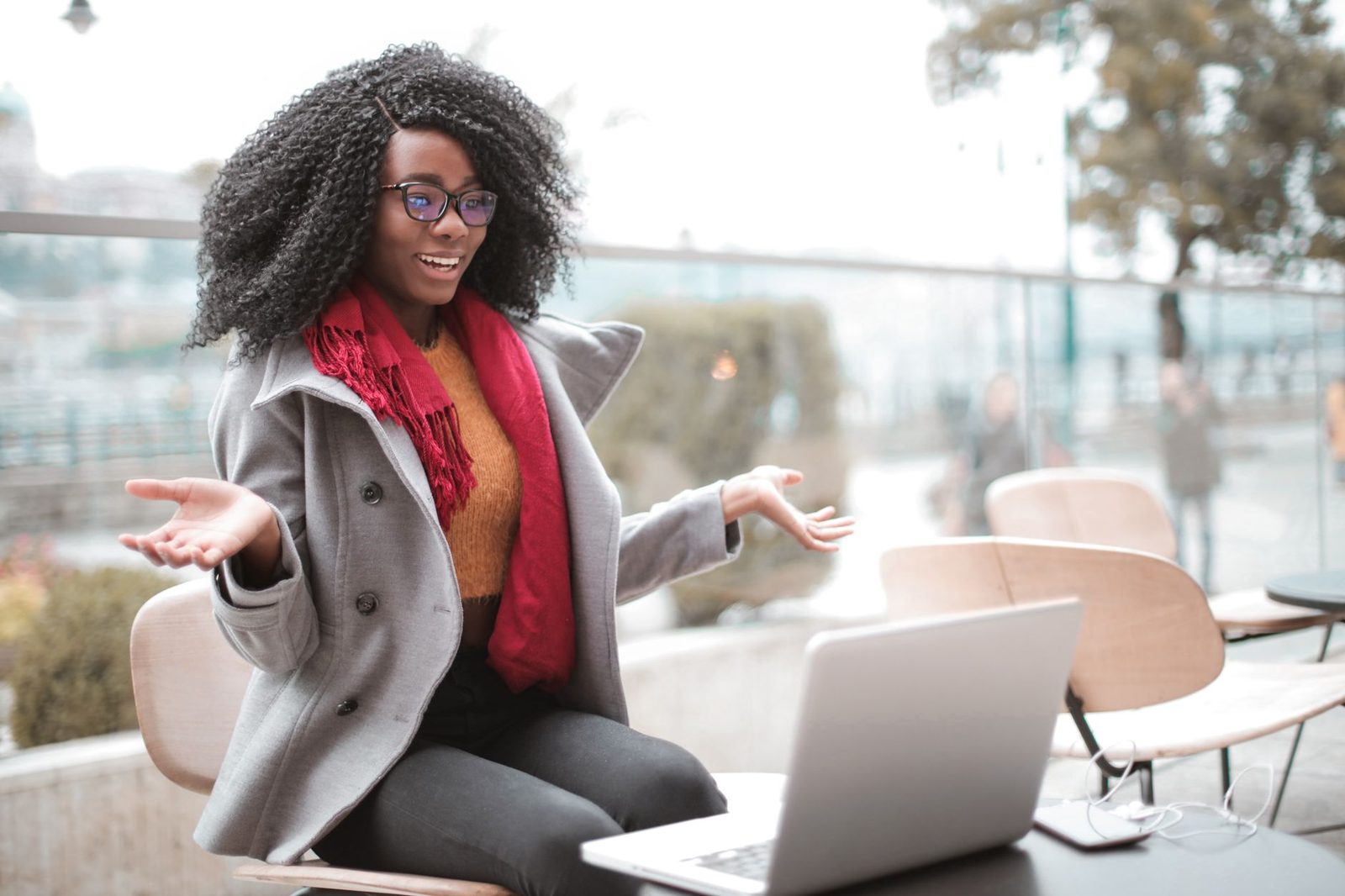 Lächelnde junge Frau mit Brille auf einer Terrasse gestikuliert vor einem Laptop