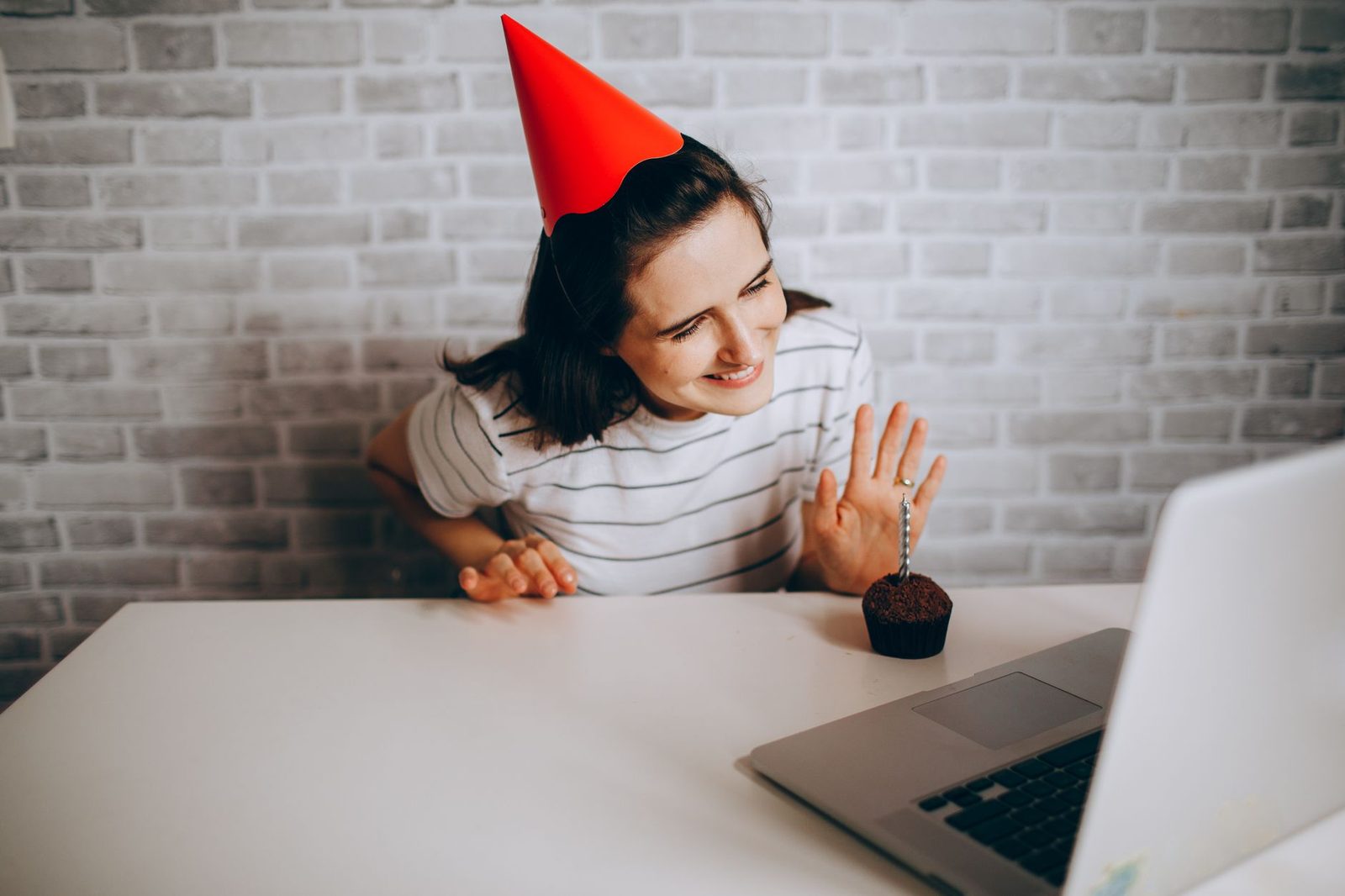 Frau mit Partyhut und Cupcake winkt vor einem Laptop-Bildschirm