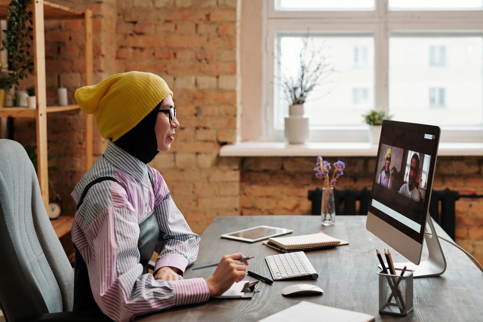 Young woman in a yellow beanie on a video call in front of a computer, two people visible on screen