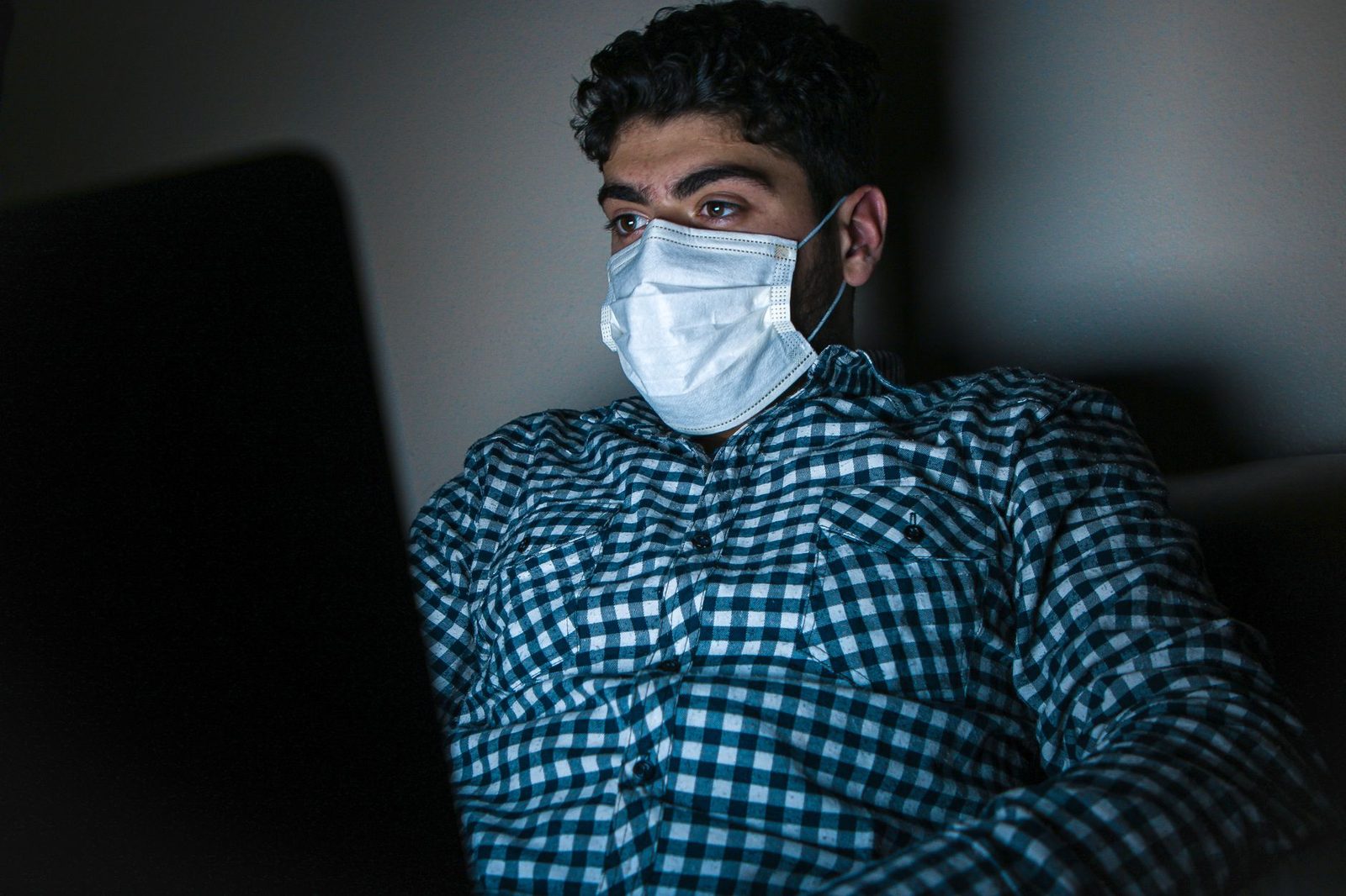 Mann mit medizinischer Maske vor einem Laptop in einem abgedunkelten Raum