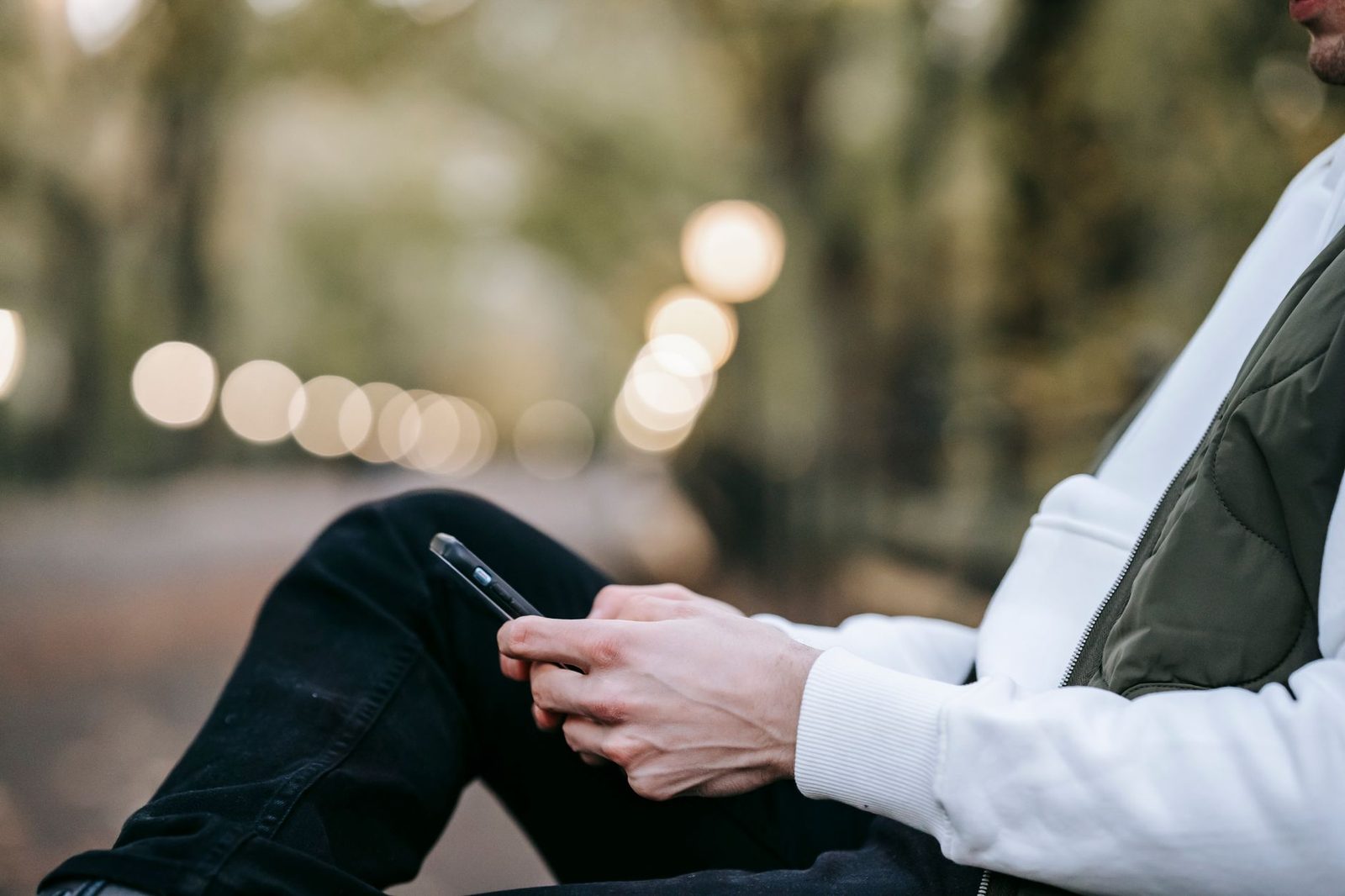 Man buiten op een bankje, in bodywarmer, met een smartphone, wazige lichten op de achtergrond