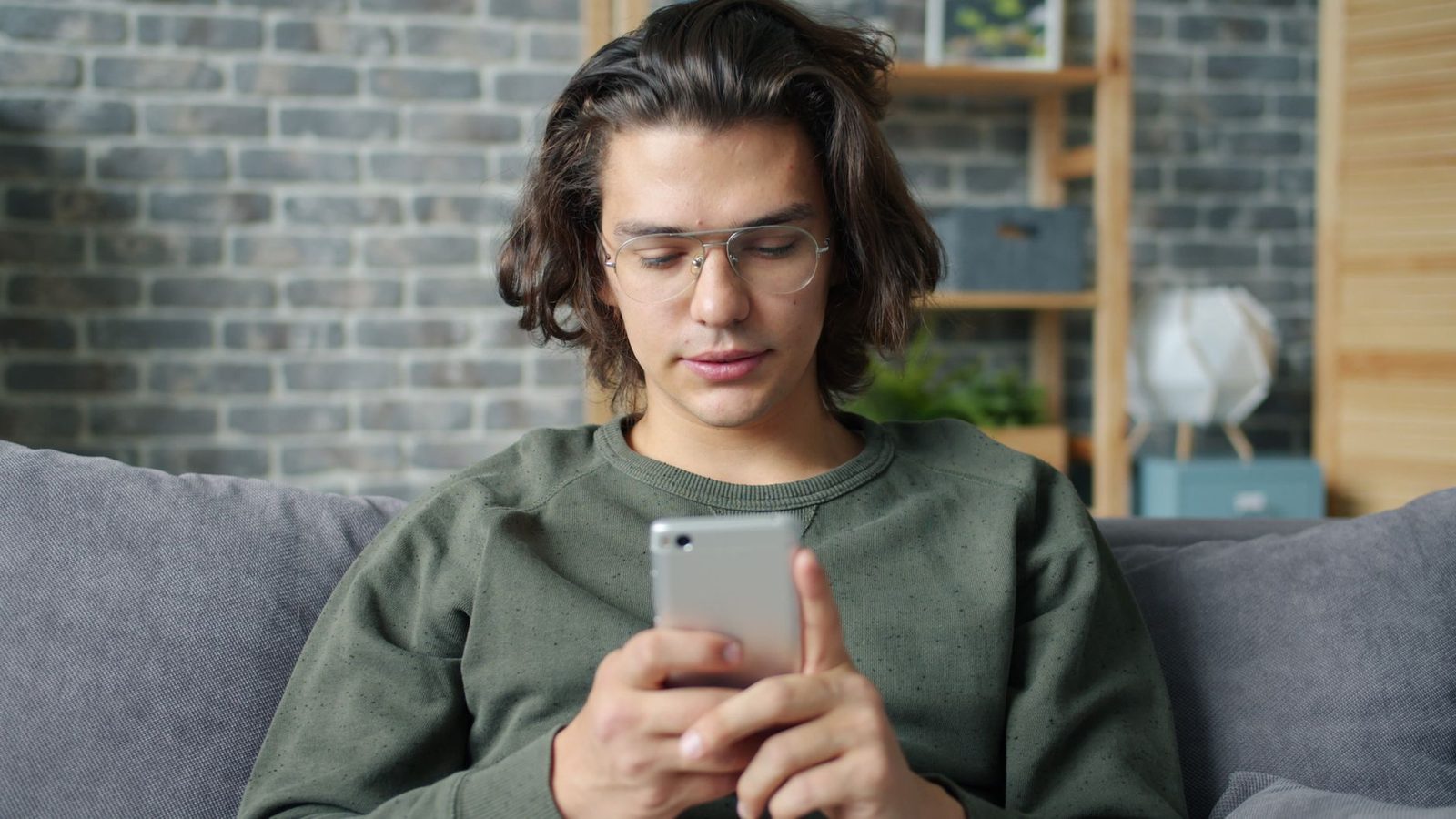 Jonge man met bril op een bank, met zijn smartphone in een woonkamer