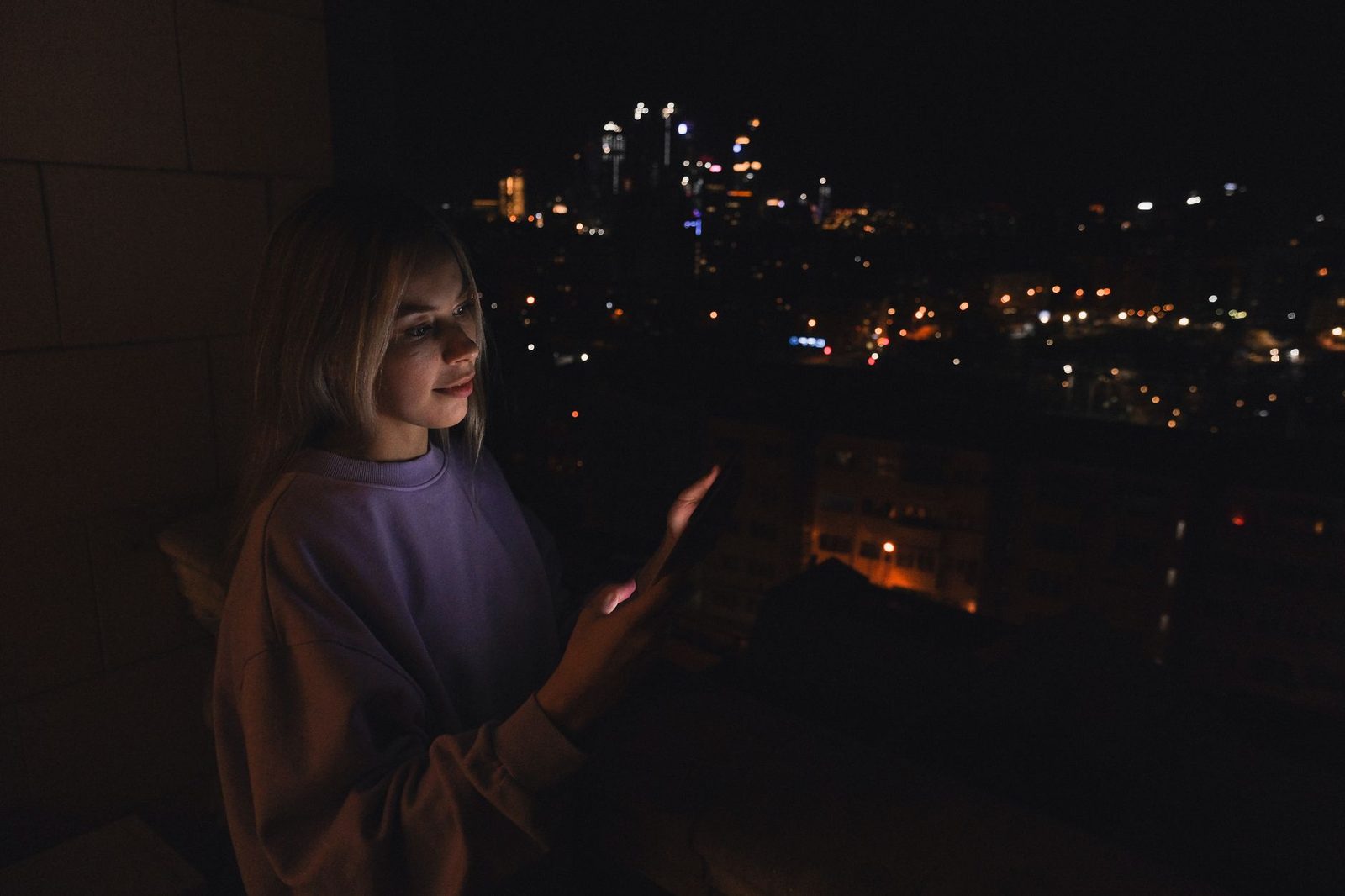 Junge Frau, die nachts auf einem Balkon ihr Smartphone benutzt, Stadtlichter im Hintergrund