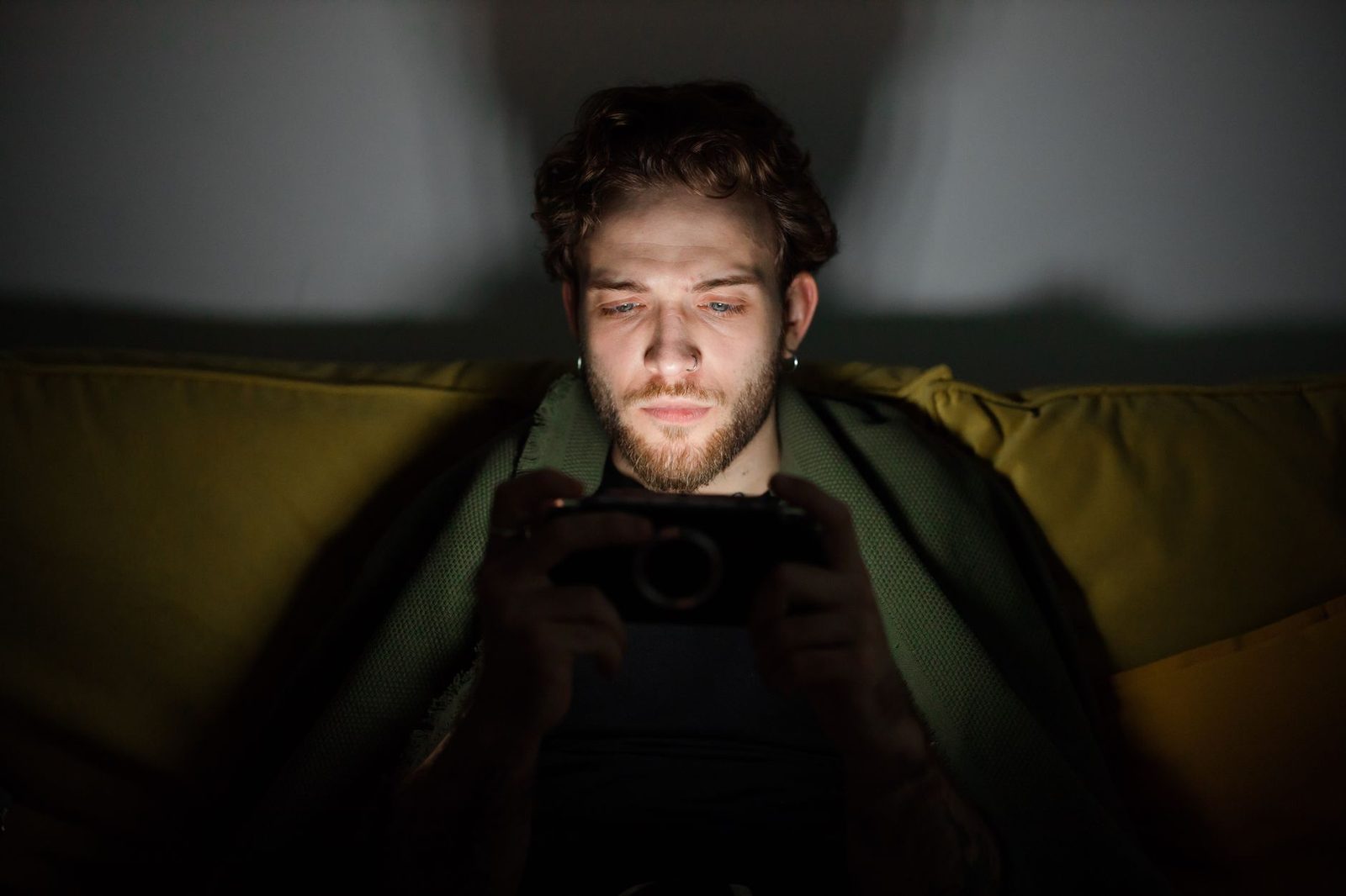 Junger Mann sitzt im Dunkeln auf einem Sofa, sein Gesicht vom Display seines Smartphones beleuchtet