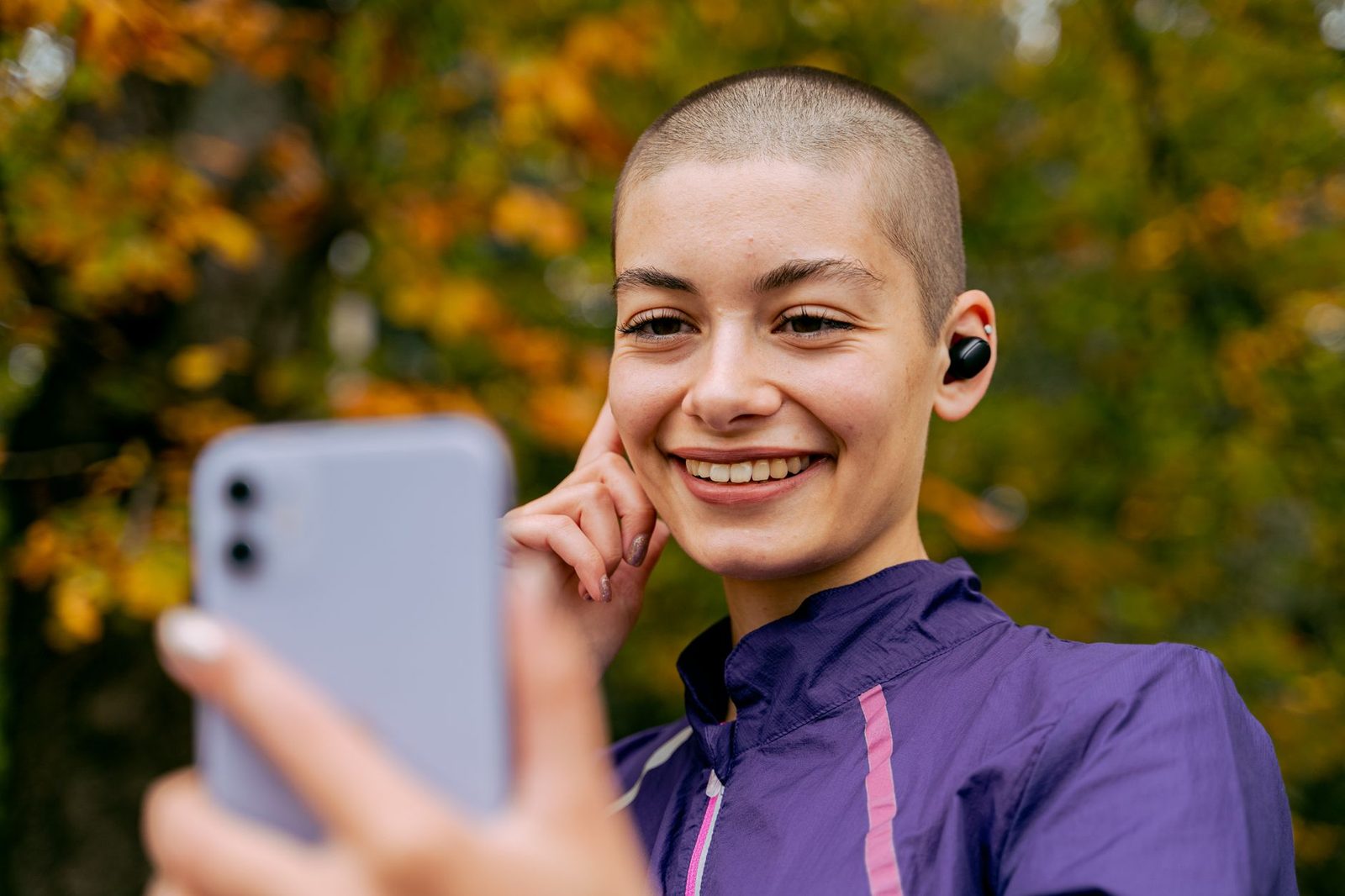 Lächelnde junge Frau mit kabellosen Kopfhörern blickt im Freien auf ihr Smartphone-Display