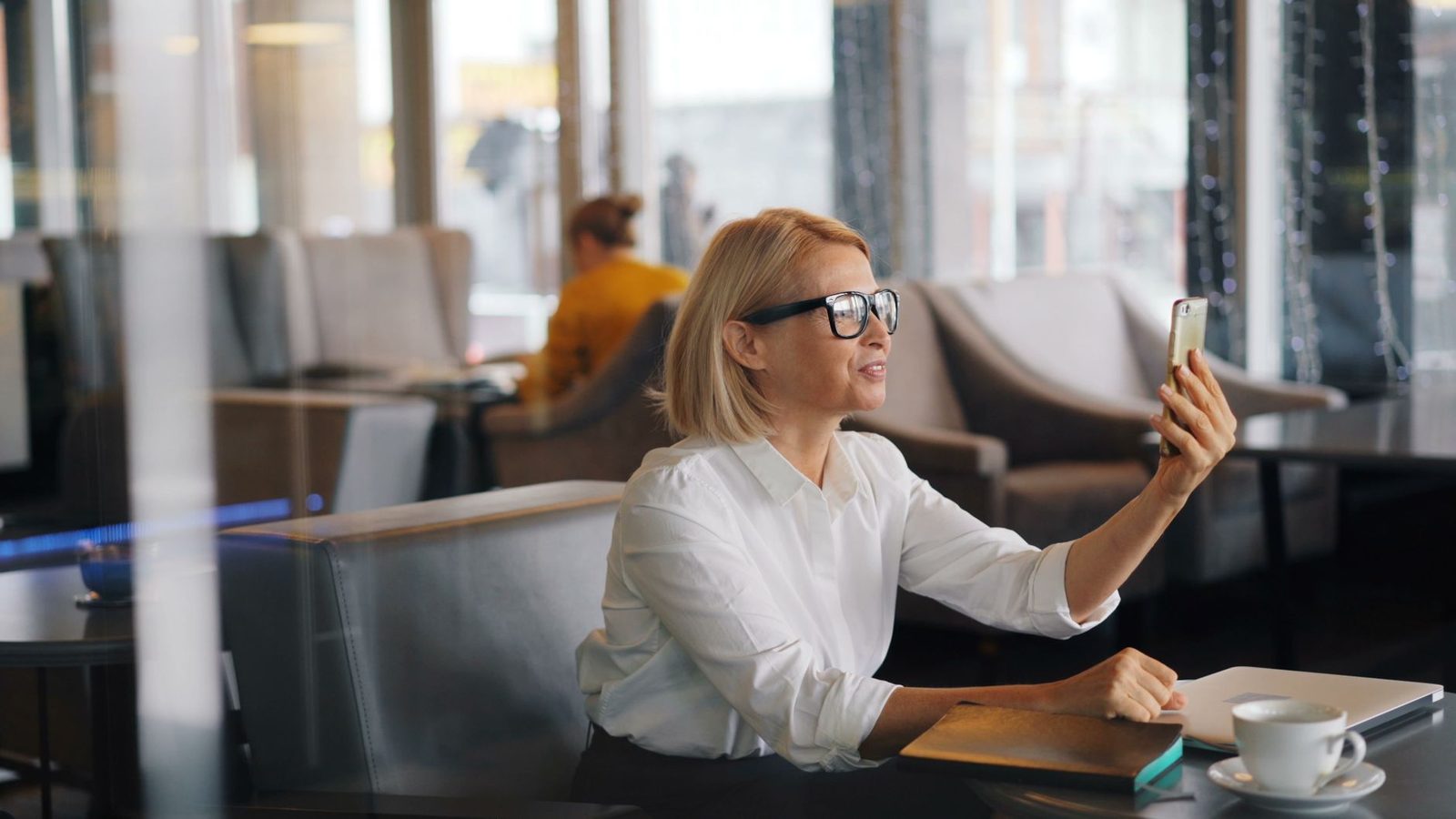 Blonde Frau mit Brille im weißen Hemd bei einem Videoanruf am Smartphone in einem Café