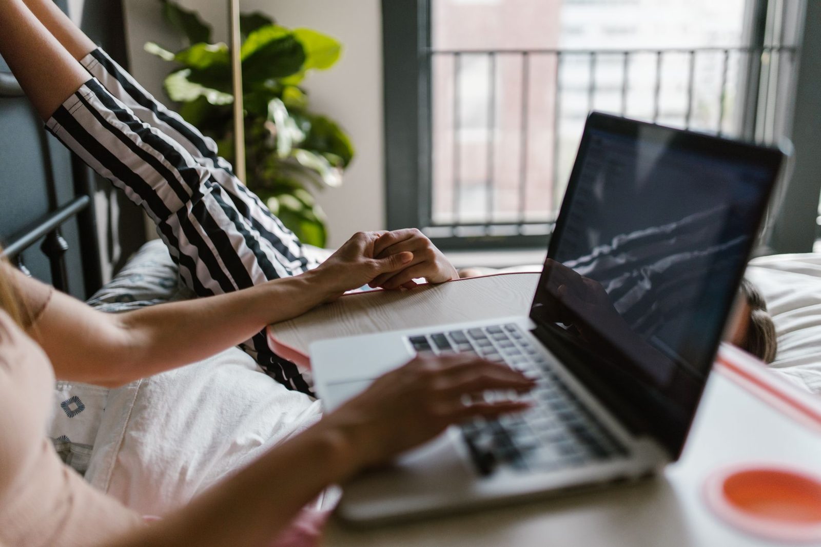 Person liegt auf einem Bett und tippt in der Nähe eines Fensters auf einem Laptop