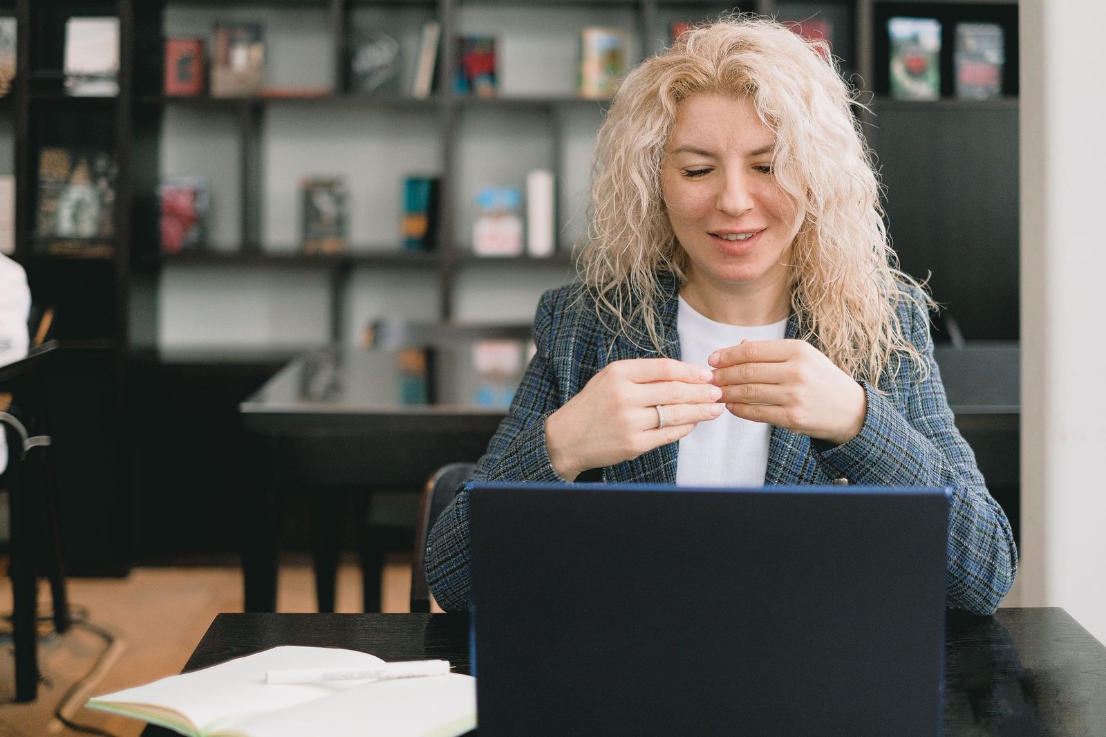Smiling blonde young woman in front of a laptop, in the middle of a video conversation in a library