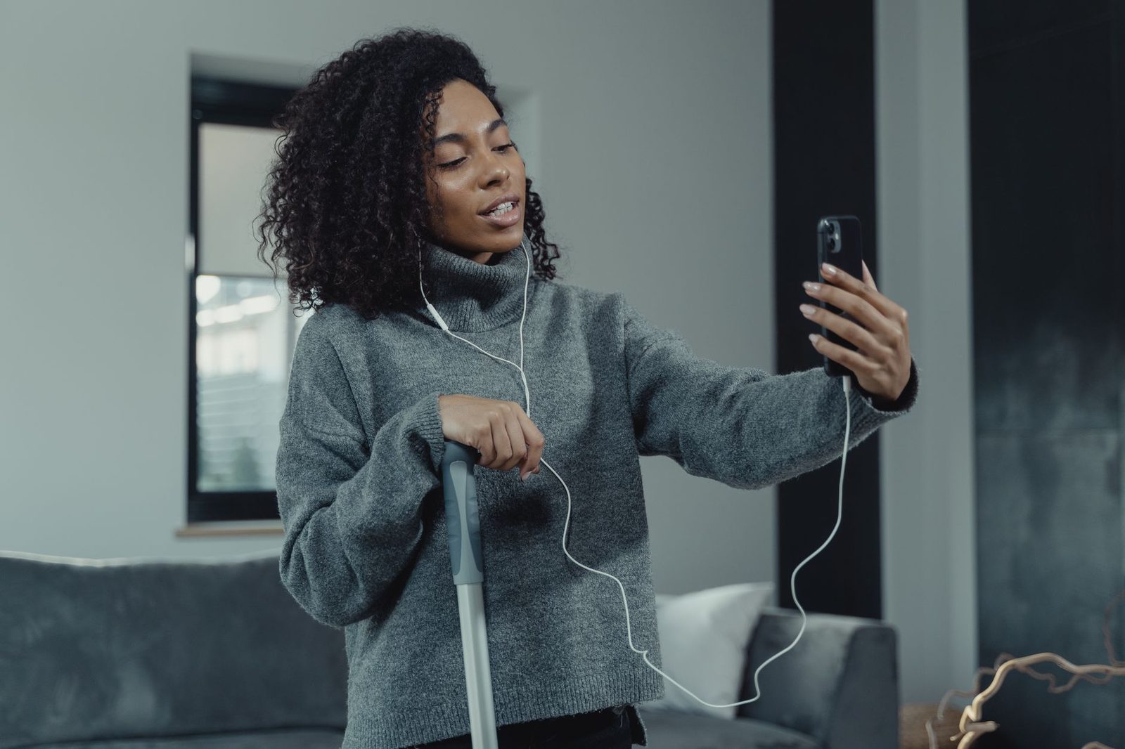Junge Frau im grauen Pullover mit kabelgebundenen Kopfhörern bei einem Videoanruf auf ihrem Smartphone im Wohnzimmer
