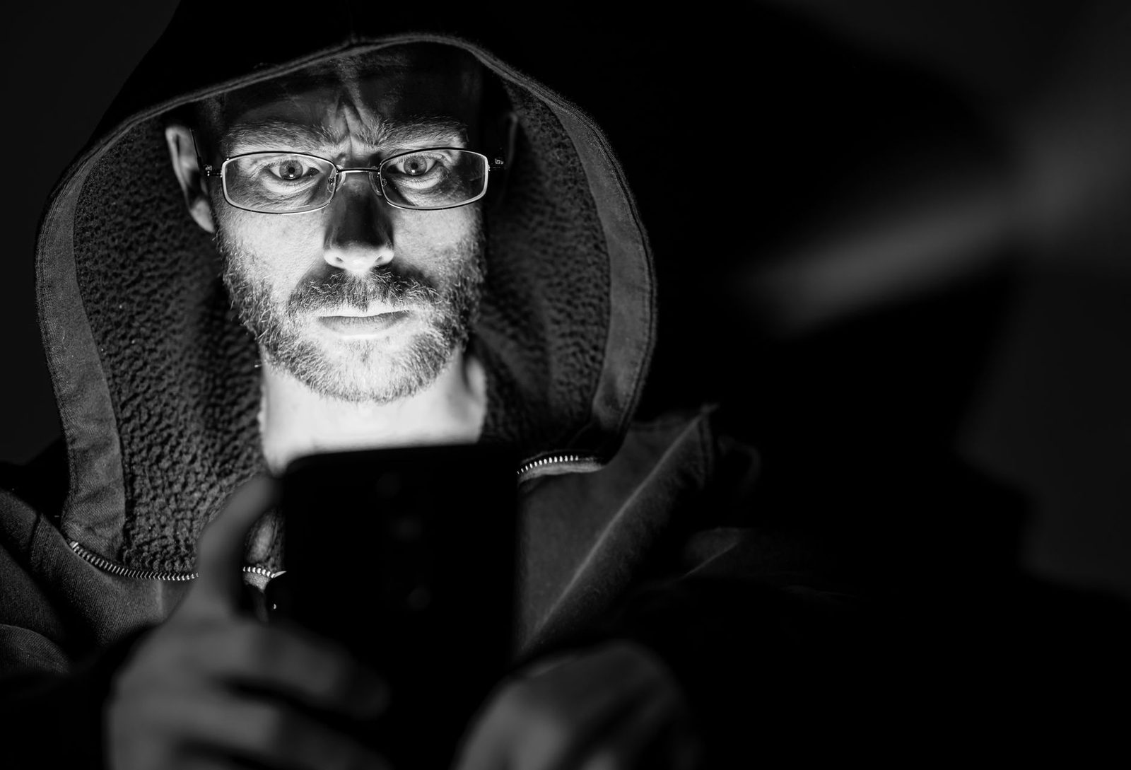 Schwarz-Weiß-Foto eines Mannes mit Brille und hochgezogener Kapuze, der im Dunkeln auf das Display seines Smartphones blickt
