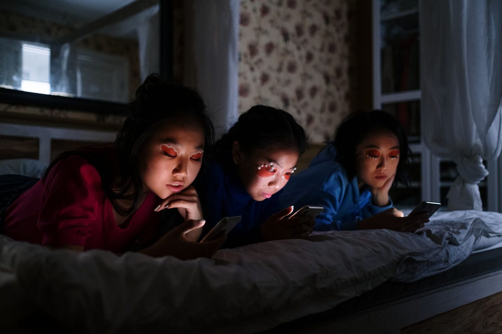 Drei junge Frauen liegen im Dunkeln auf einem Bett und schauen auf das leuchtende Display ihres Smartphones