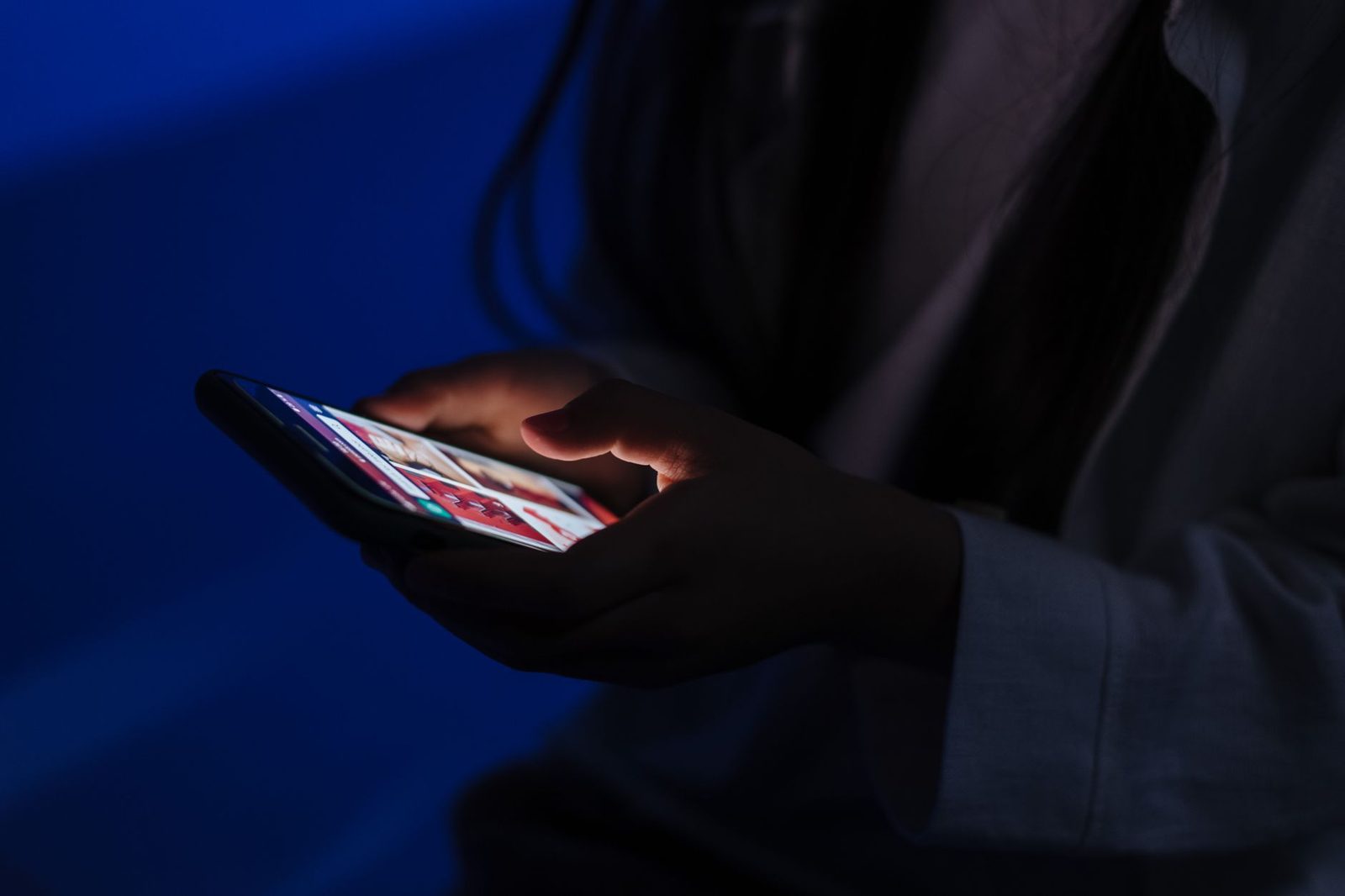 Hände einer jungen Frau, die ein eingeschaltetes Smartphone in einem dunklen, blau beleuchteten Raum hält