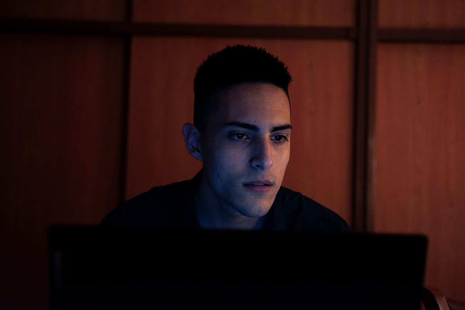 Jonge man met zijn gezicht verlicht door het scherm van een laptop in een donkere kamer