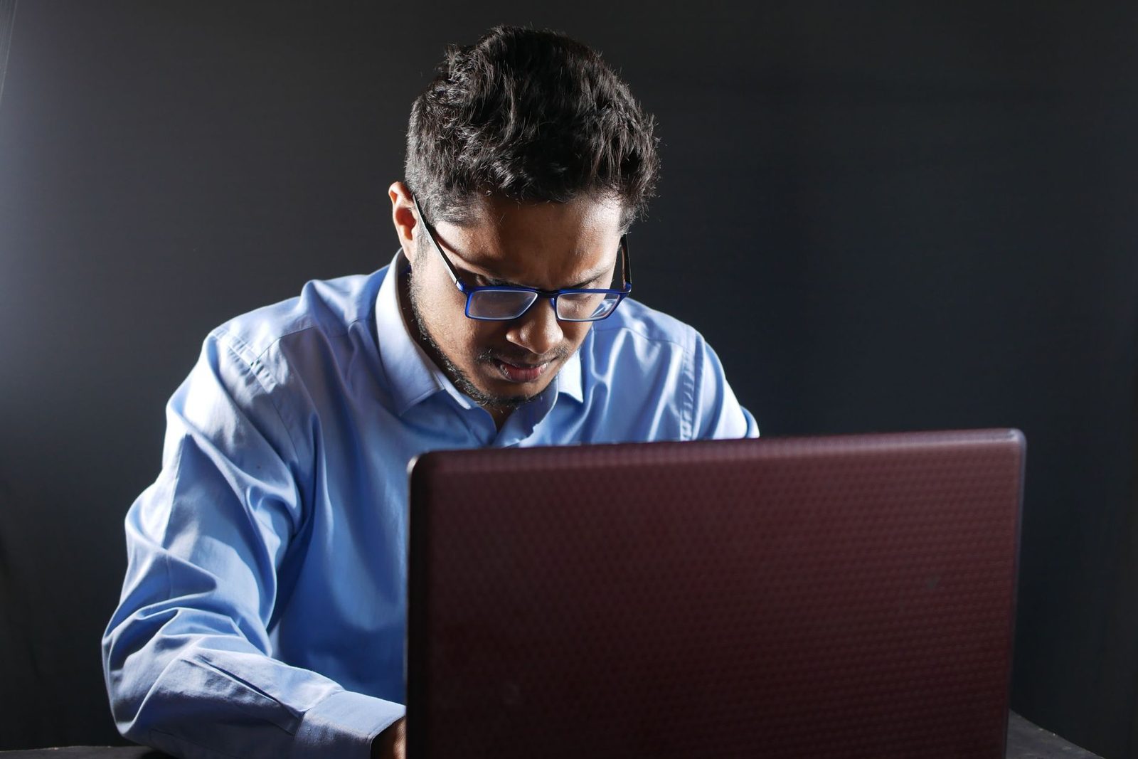 Man met bril in een blauw overhemd voor een laptop, tegen een zwarte achtergrond