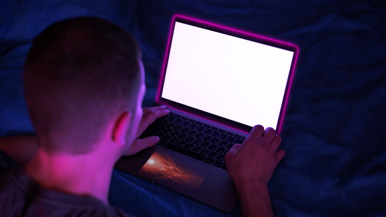 Mann liegt im Dunkeln vor einem Laptop mit weißem Bildschirm, angestrahlt von einem rosafarbenen Schimmer