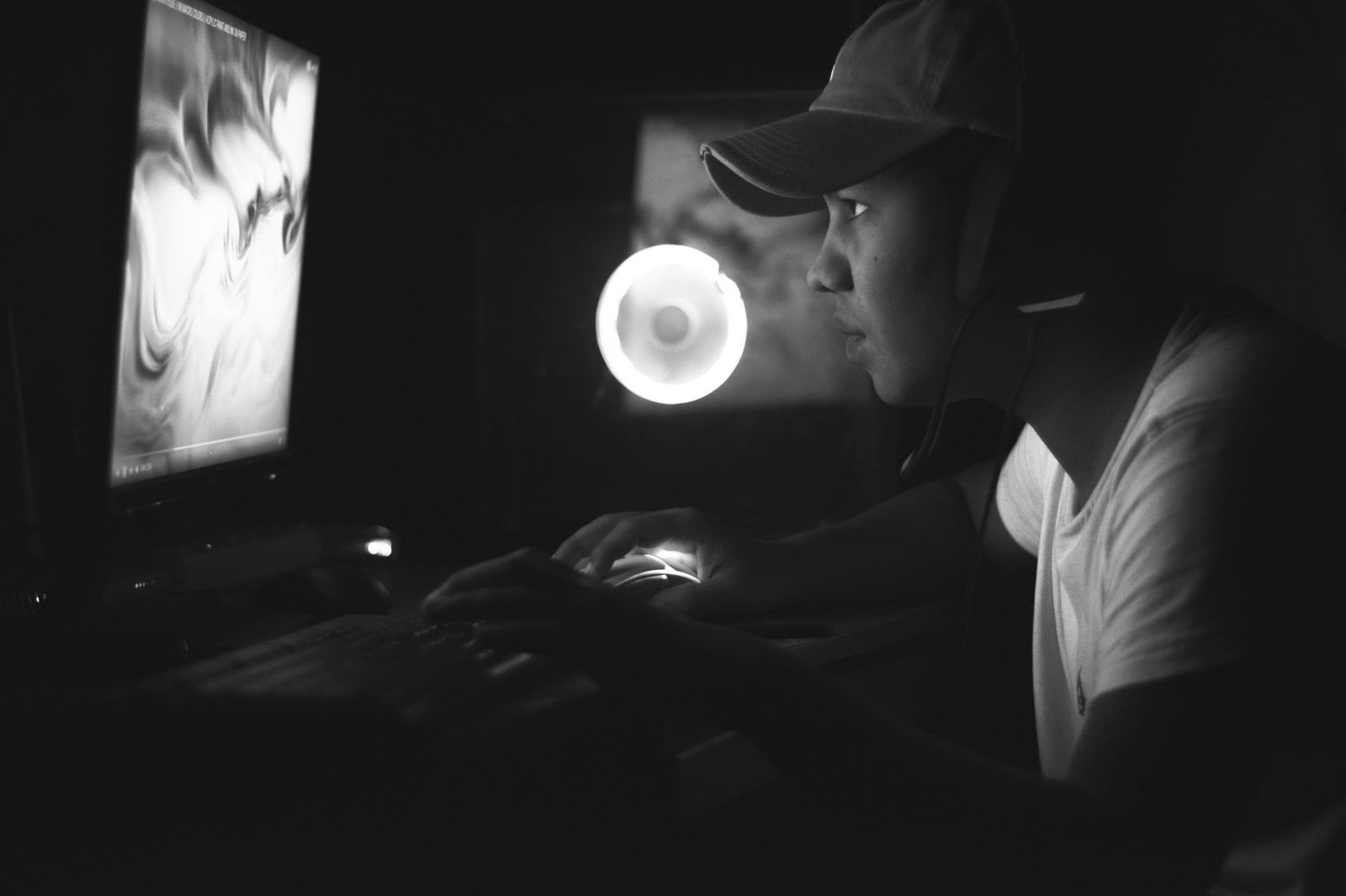 Schwarz-Weiß-Foto eines jungen Mannes mit Cap und Headset vor einem Computerbildschirm im Dunkeln