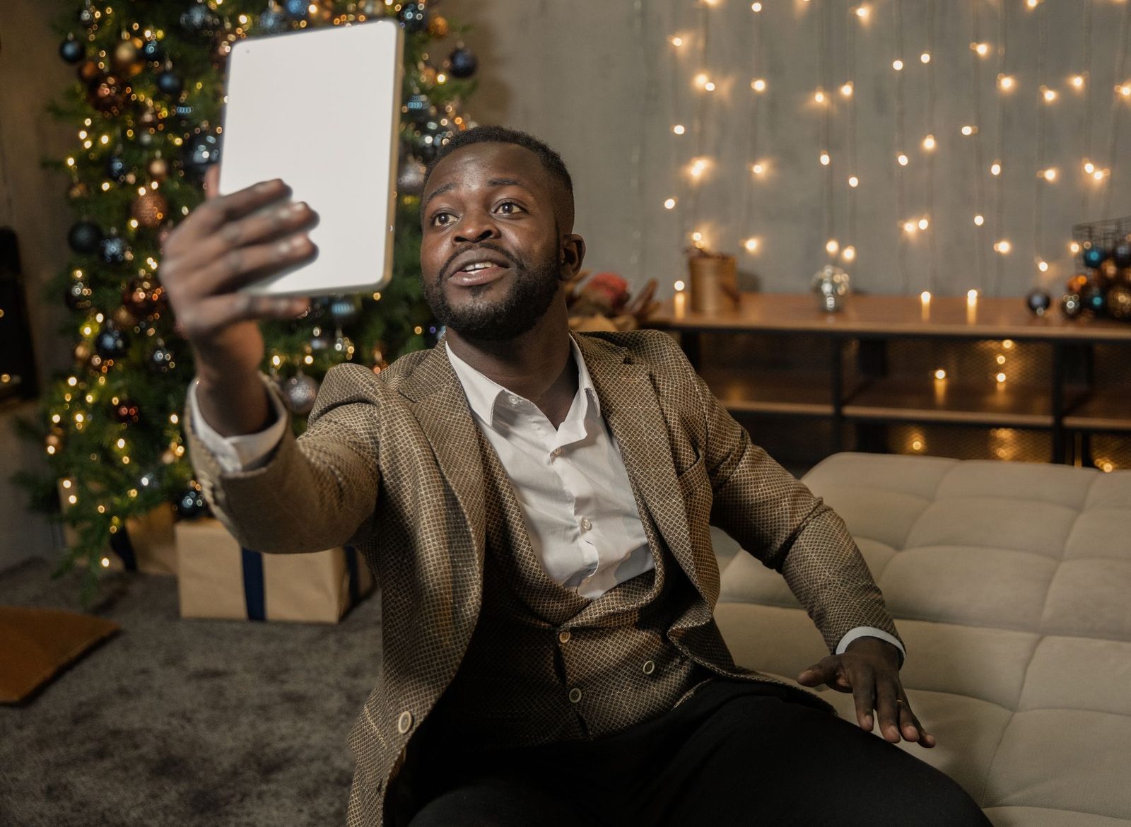 Mann in Jacke hält ein Tablet mit ausgestrecktem Arm für einen Videoanruf, vor einem Tannenbaum und Lichterketten